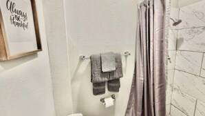 Een badkamer met douchegordijn, een handdoekrek met twee handdoeken, een lijst met een citaat aan de wand en een wc-rolhouder.