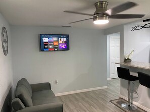 A living room with a flat-screen TV mounted on the wall, a ceiling fan, a grey sofa, and a kitchen area with a bar stool.