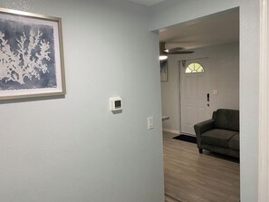 Un pasillo con una obra de arte enmarcada, un termostato y una puerta que da a una habitación con sofá.