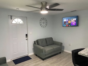A living room with a gray sofa, a ceiling fan, a mounted TV, and a wall clock.