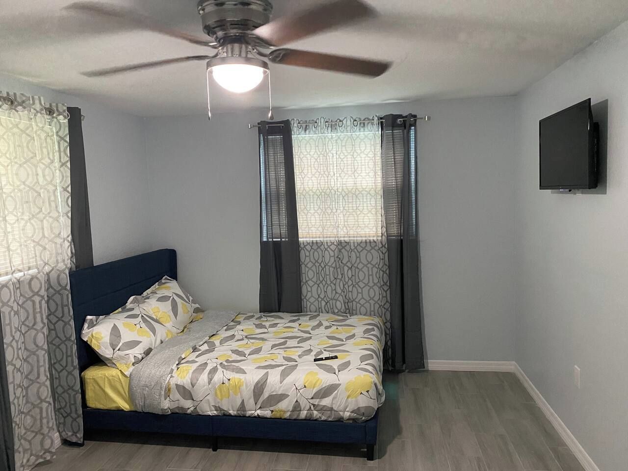 Une chambre à coucher avec un lit, des rideaux, un ventilateur de plafond et un téléviseur fixé au mur.