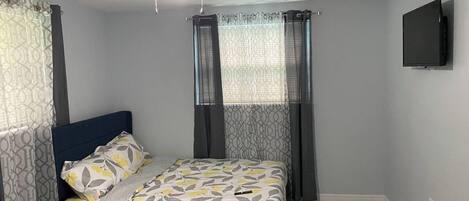 Une chambre à coucher avec un lit, des rideaux, un ventilateur de plafond et un téléviseur fixé au mur.