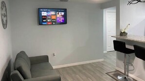 A modern living room with a grey sofa, a flat-screen TV mounted on the wall, a ceiling fan, and a kitchen area with a bar stool. - Modern 2/1 Sleeps 6 Quiet Dead End St 1/2 Acre Wow (Parrish)