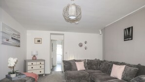 Ein Wohnzimmer mit grauem Ecksofa, einem grauen Hocker, einer weißen Lampe und einer Lampenschirm aus Weidengeflecht.