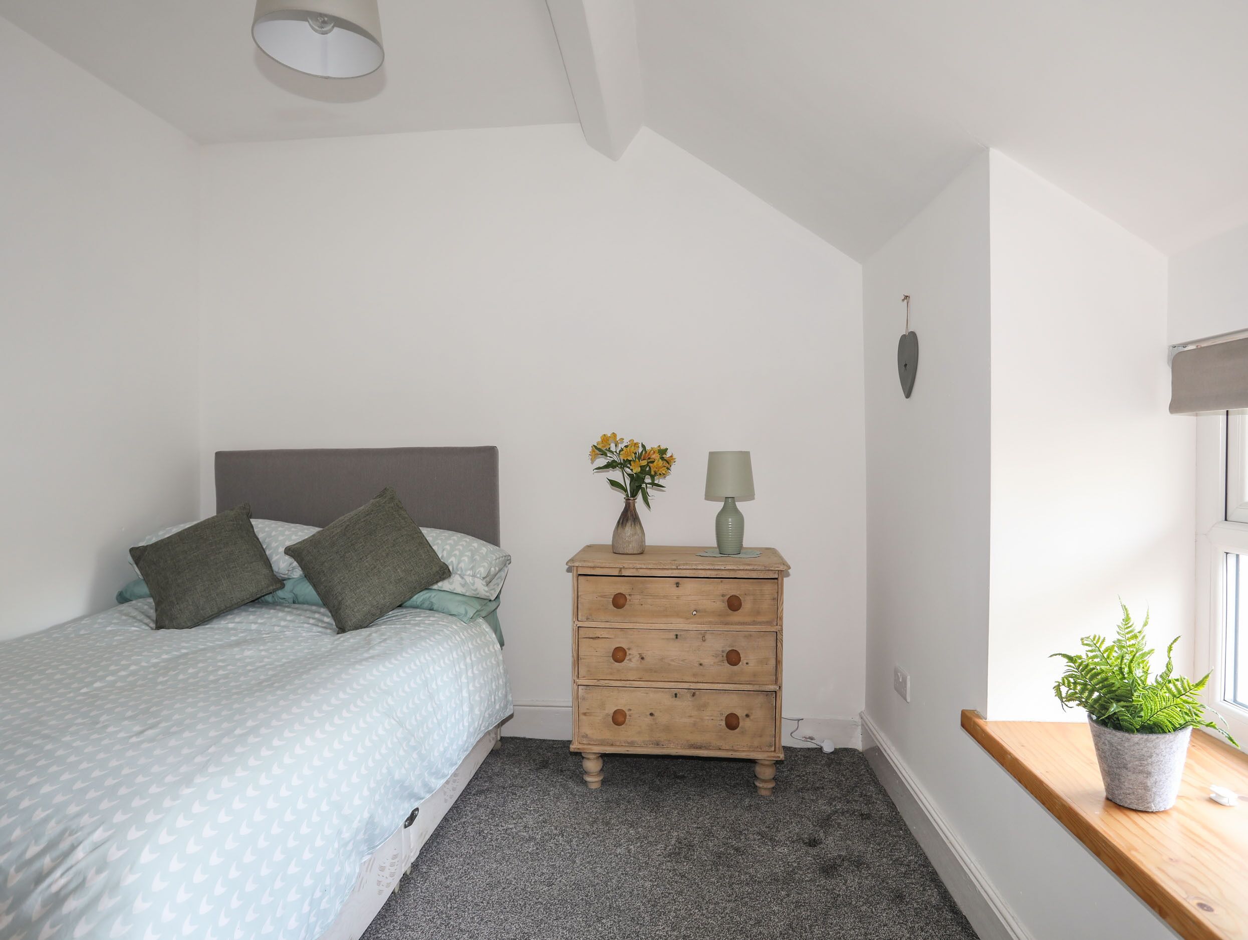 Un dormitorio con cama, cómoda de madera, una mesita con planta y ventana con planta en el alféizar.