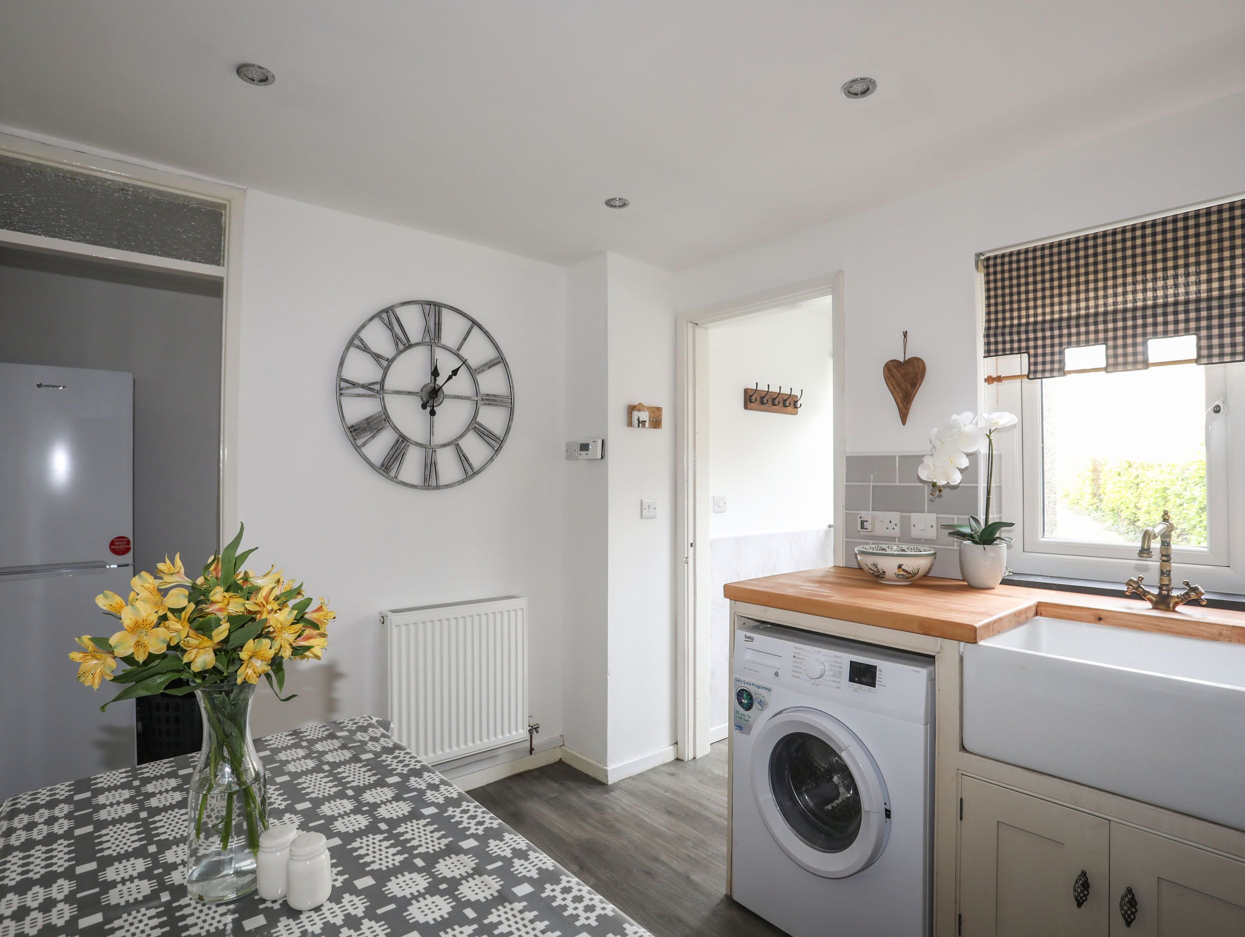 A modern kitchen with a built-in washing machine, a large wall clock, and a window with a checkered valance.