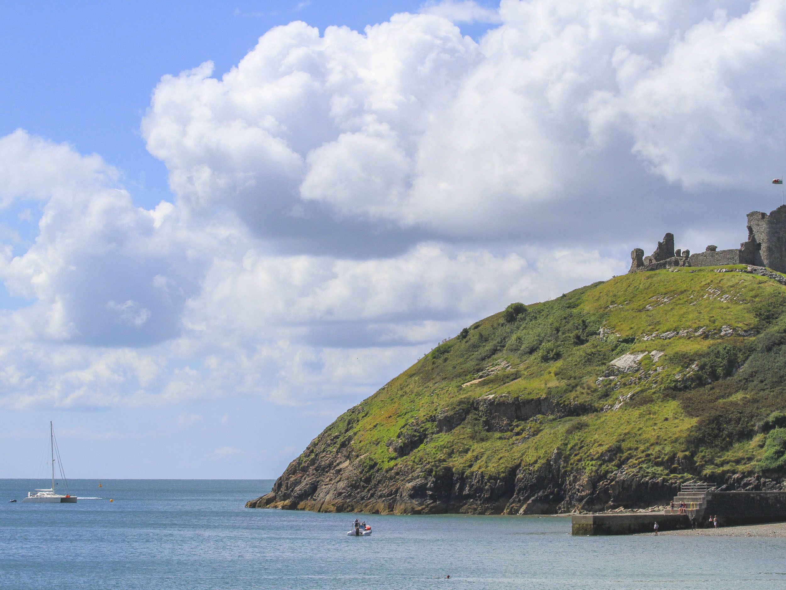 A coastal scene with a castle on a hill, calm sea, and a sailboat.