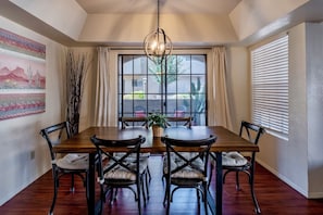 Une salle à manger avec une table et des chaises en bois, un luminaire suspendu, une fenêtre avec des stores et un tableau au mur.