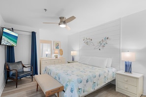 A bedroom with a bed, a desk, a chair, a TV, a ceiling fan, and a nightstand with lamps.
