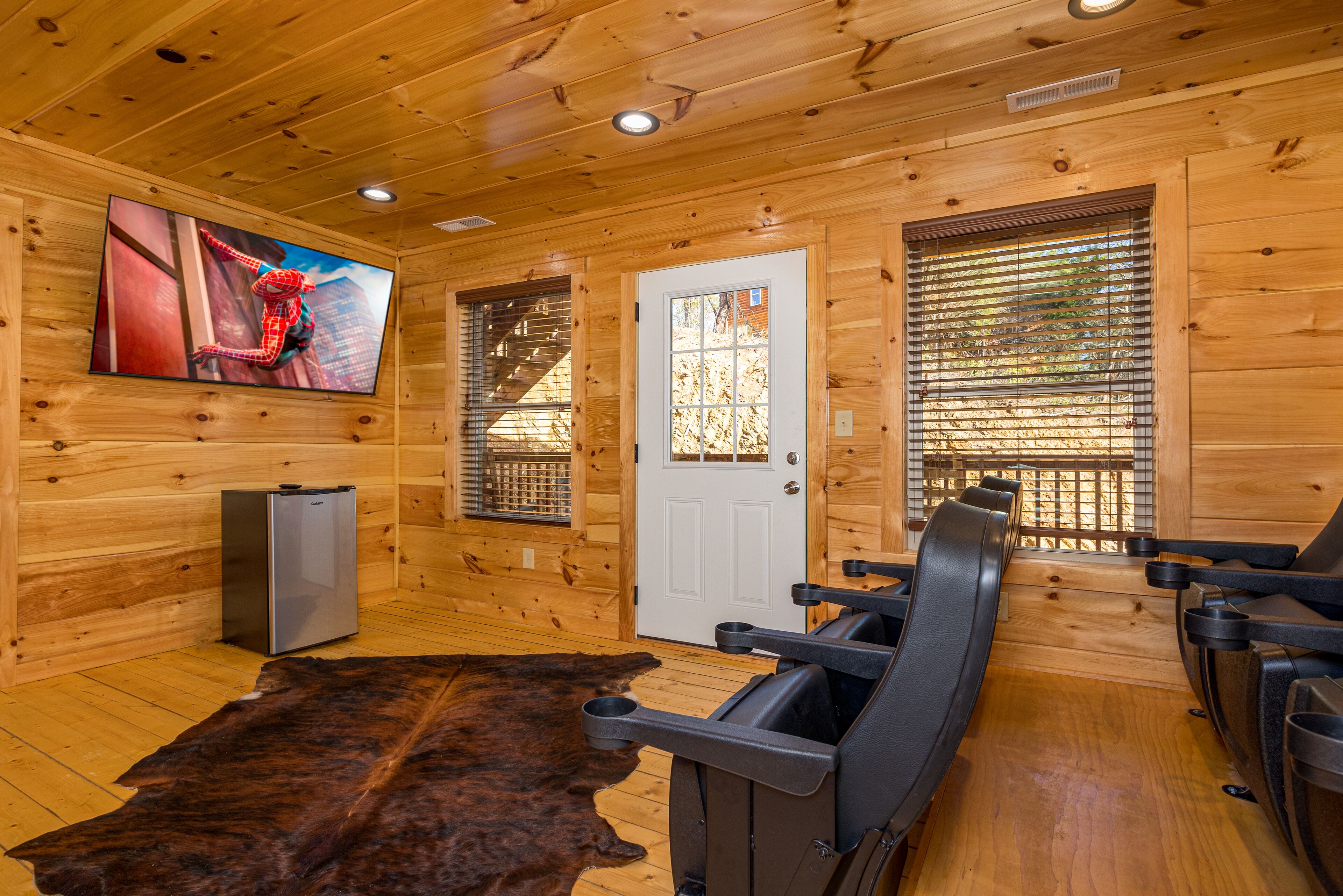 Interior de una cabaña de madera con una televisión grande, un refrigerador y un sofá de cuero.