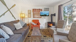 A cozy living room with a sofa, armchairs, a wooden coffee table, a fireplace, and a large window with a view of trees. - Family Lakefront Cabin! Hot Tub, Arcades & Dock (Wautoma)