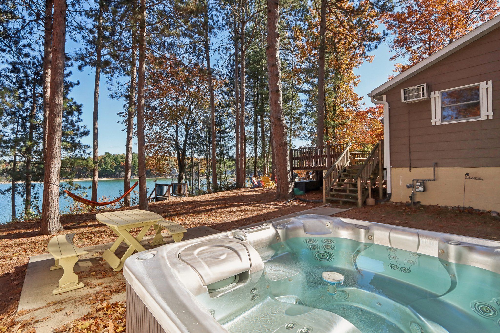 A hot tub in a backyard with a wooden deck, a hammock, and a view of a lake.