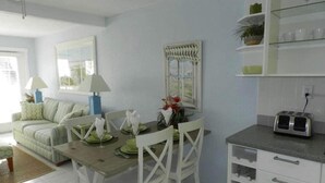 Un comedor con una mesa de madera dispuesta para cuatro, sillas blancas, una mesita auxiliar con una lámpara y una ventana con vistas.