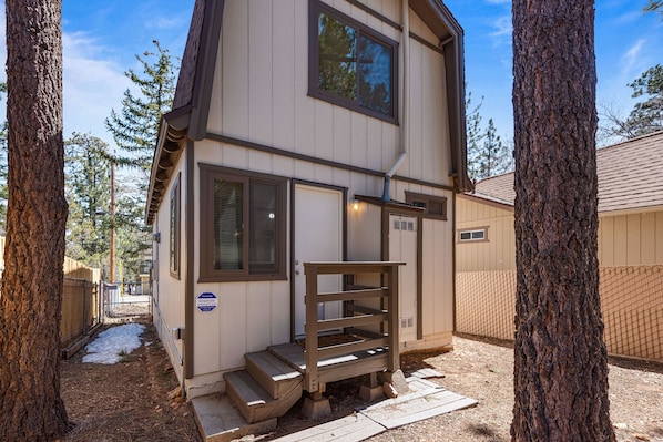 A two-story cabin with a wooden staircase leading to the entrance, surrounded by trees and a fenced yard.