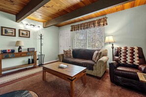 Una sala de estar acogedora con techo de madera, un sofá, un sillón de cuero, una mesita de centro y una mesita auxiliar con una lámpara.