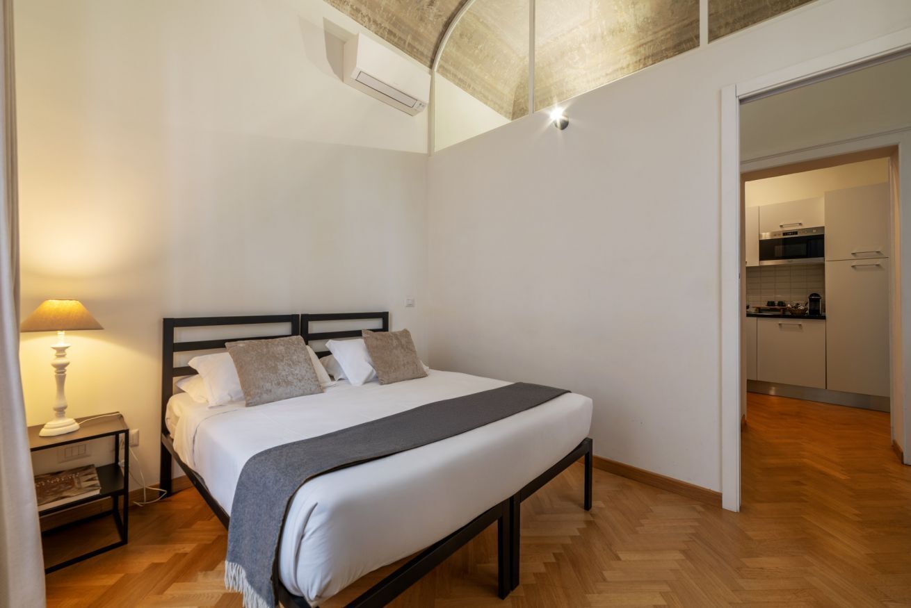 Ein Schlafzimmer mit Bett, Nachttisch, Lampe und Blick auf die Küche.
