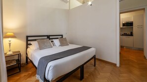 A bedroom with a bed, bedside table, lamp, and a view of the kitchen. - Castro Pretorio 3 - FromHometoRome (Roma)