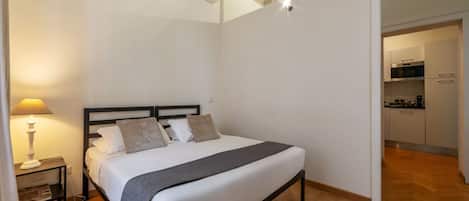 A bedroom with a bed, bedside table, lamp, and a view of the kitchen.