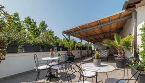 An outdoor patio with round tables and chairs, surrounded by potted plants and a pergola.