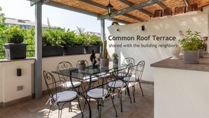A common roof terrace with a glass table and chairs, surrounded by potted plants and a thatched roof. - Castro Pretorio 3 - FromHometoRome (Roma)