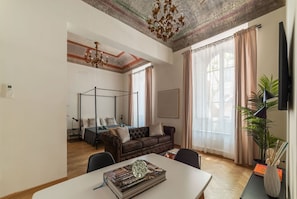 A modern living room with a large window, a brown leather sofa, a dining table with a decorative book, and a ceiling with exposed beams and a chandelier.
