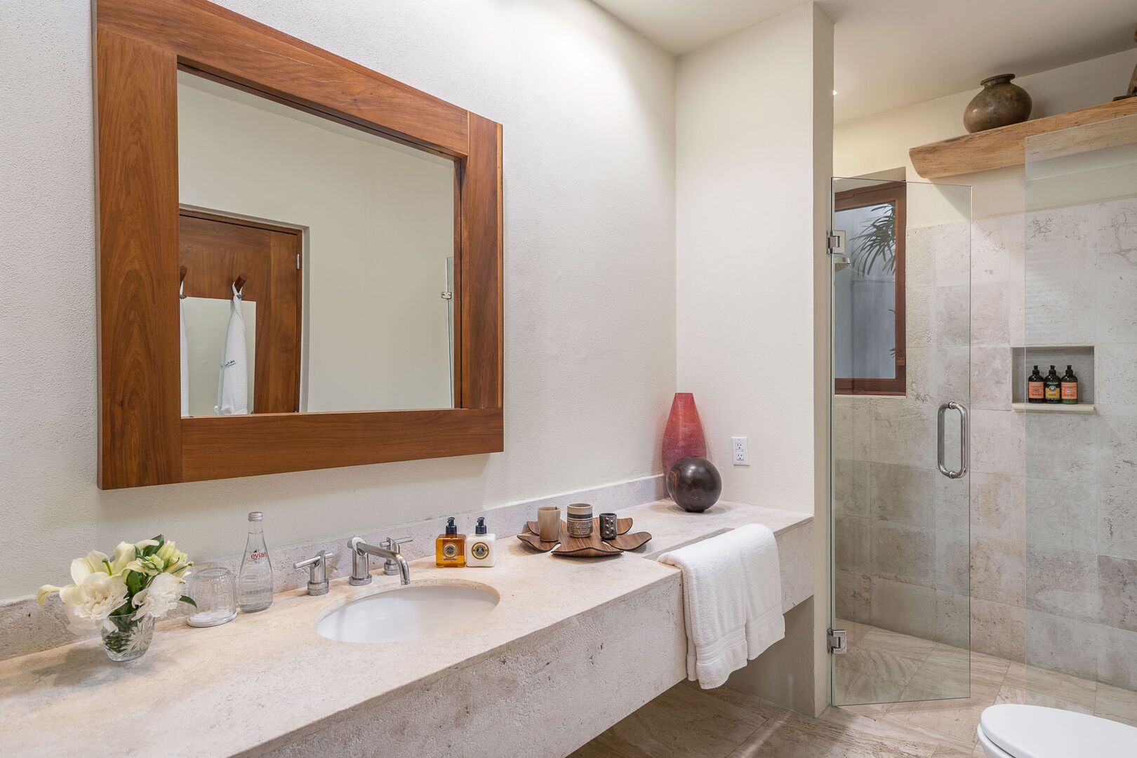 Un baño moderno con un espejo de marco de madera, un lavabo con artículos de aseo, una zona de ducha y un inodoro.