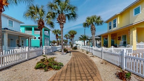 A pathway lined with palm trees and a white picket fence, leading to a row of colorful beach houses. - Beautiful Home in Gated Village Walk, 2 Shared Heated Pools, Pets Allowed, Garage Access (Port Aransas)