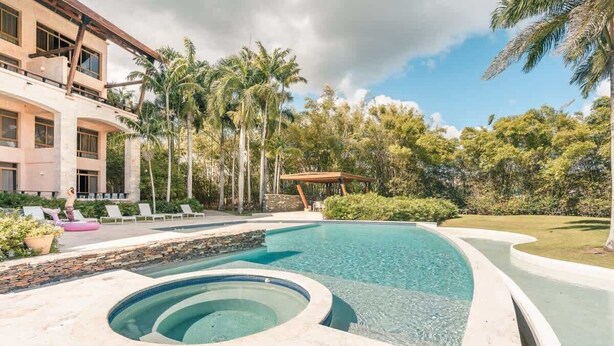A swimming pool with a hot tub, lounge chairs, and a covered pavilion surrounded by palm trees and greenery. - Luxury Golf Front Apartment with Pool Access (Casa De Campo)
