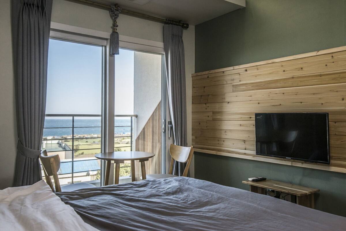A hotel room with a large bed, a small table, two chairs, a TV mounted on a wooden wall, and a balcony with a view of the sea.