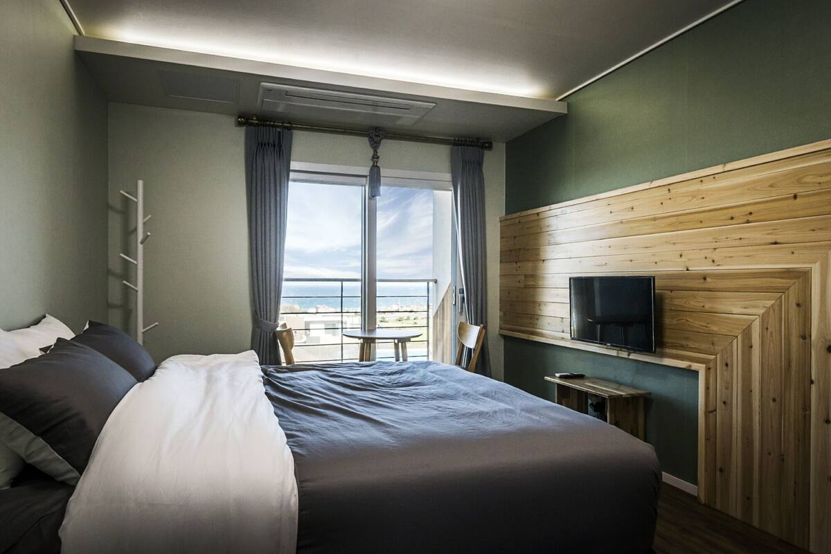 A hotel room with a bed, a TV mounted on a wooden wall, a balcony with a view, and a chair.