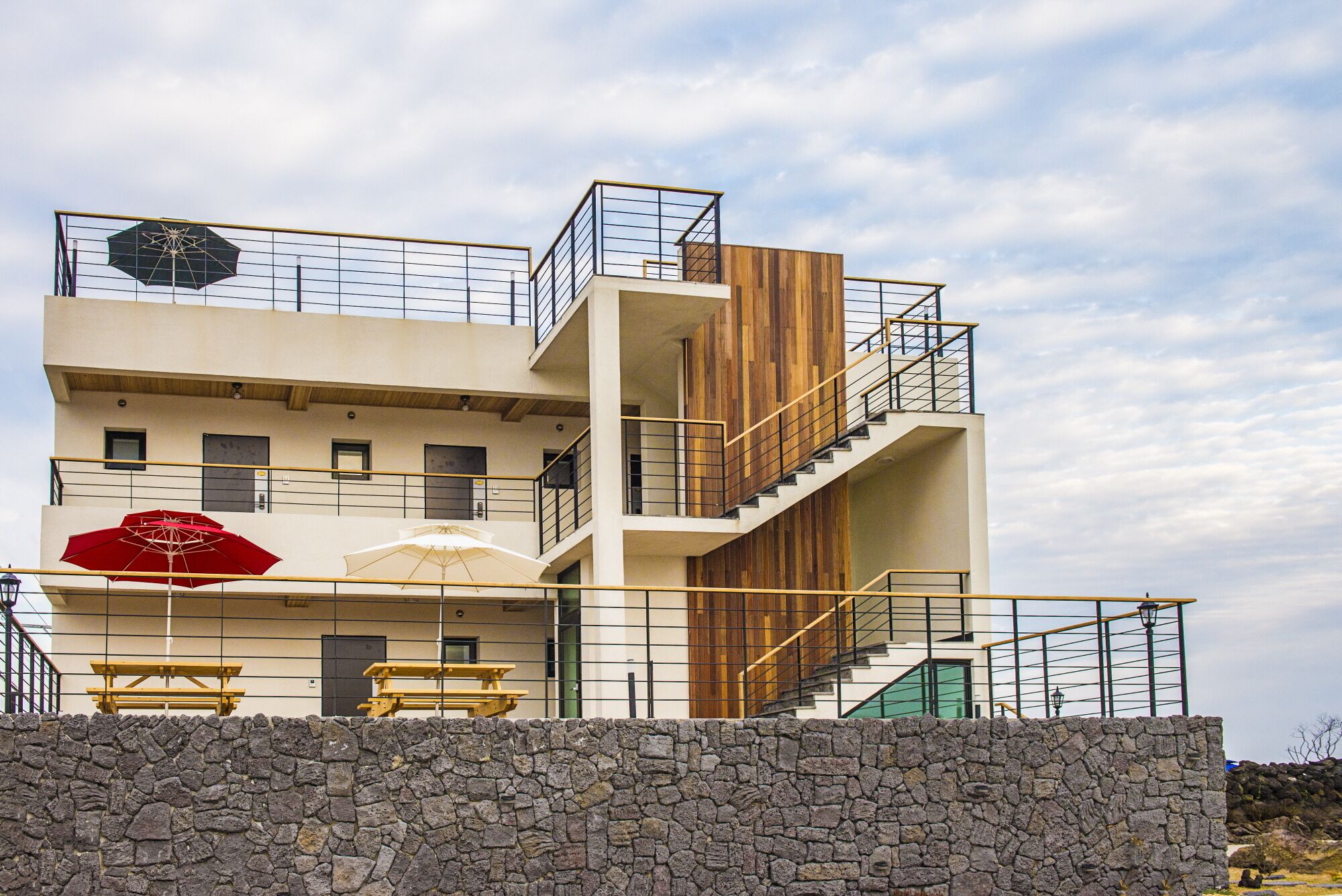 Un edificio de varios pisos con un muro de piedra en primer plano, paneles de madera en los pisos superiores y un paraguas rojo en la planta baja.
