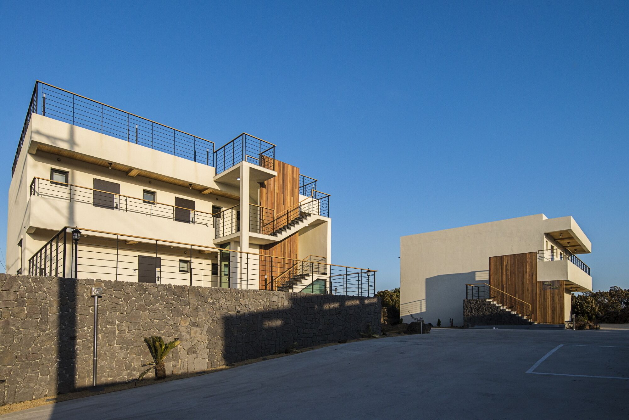 A modern multi-story building with balconies, a stone wall, and a parking area.
