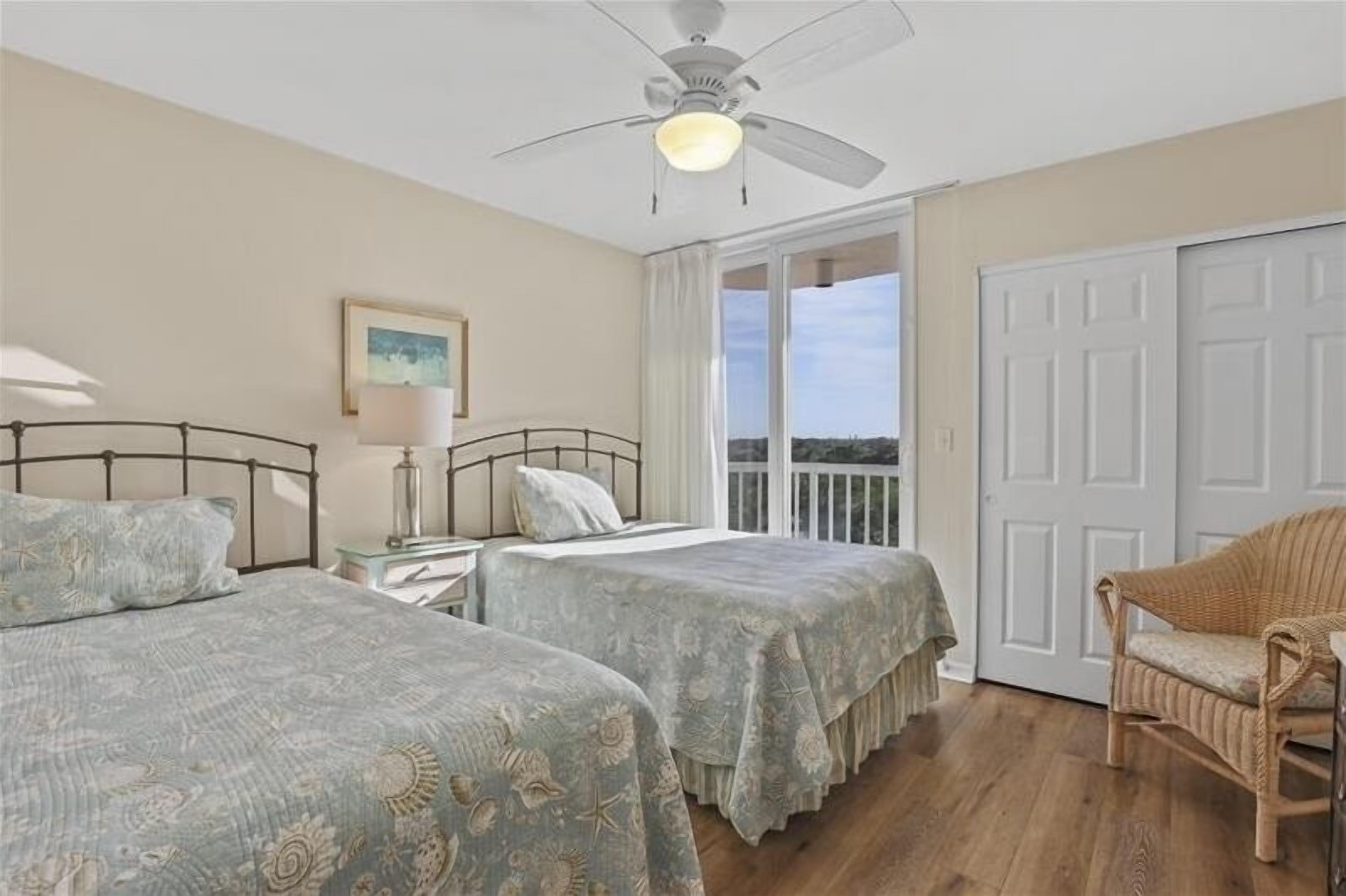 A bedroom with two beds, a ceiling fan, a window with a view, and a wicker chair.