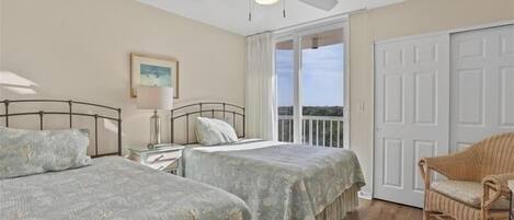 A bedroom with two beds, a ceiling fan, a window with a view, and a wicker chair.