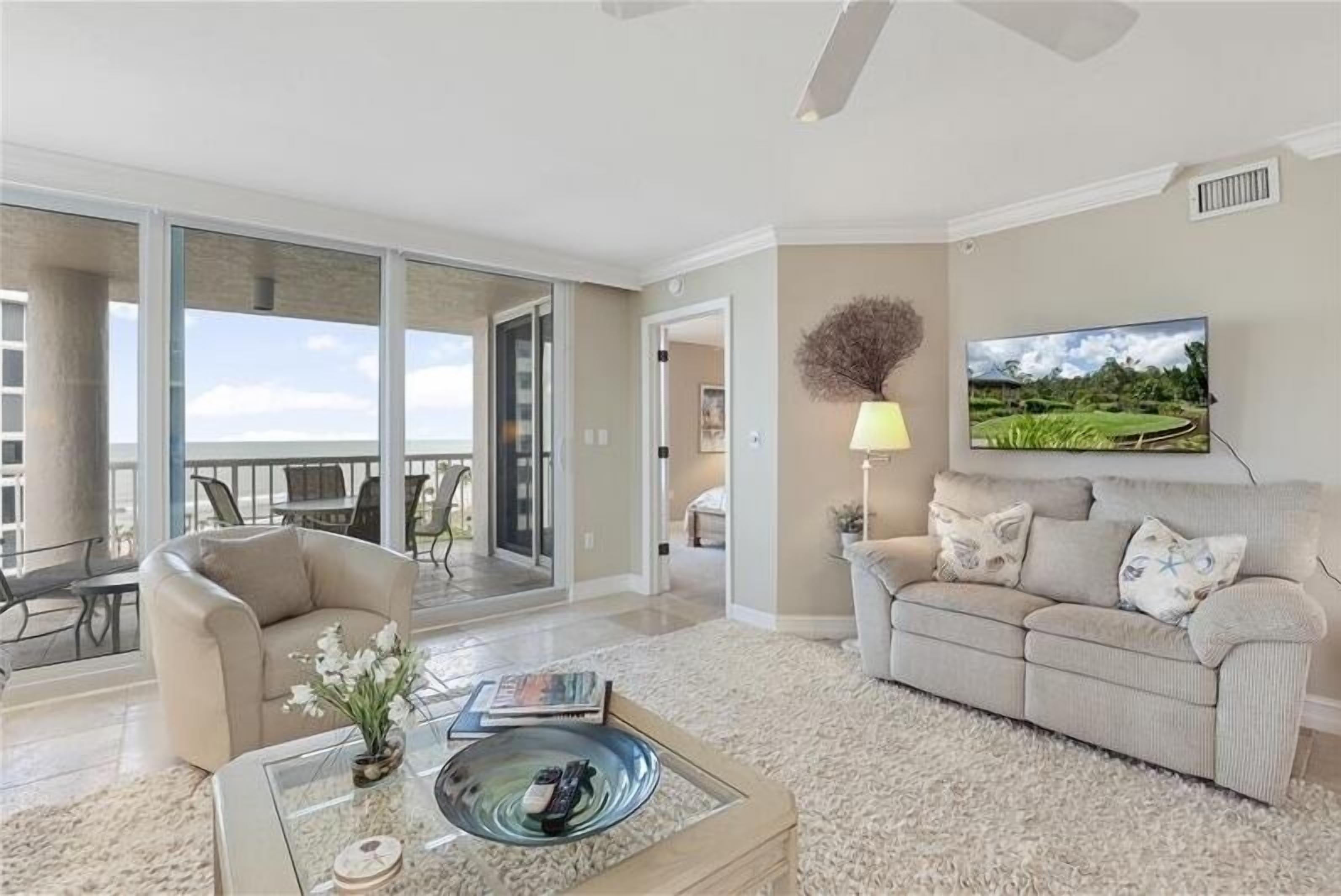 A spacious living room with a large sliding glass door leading to a balcony, a comfortable sofa, a coffee table with remote controls, and a television mounted on the wall.