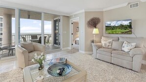 A spacious living room with a large sliding glass door leading to a balcony, a comfortable sofa, a coffee table with remote controls, and a television mounted on the wall.
