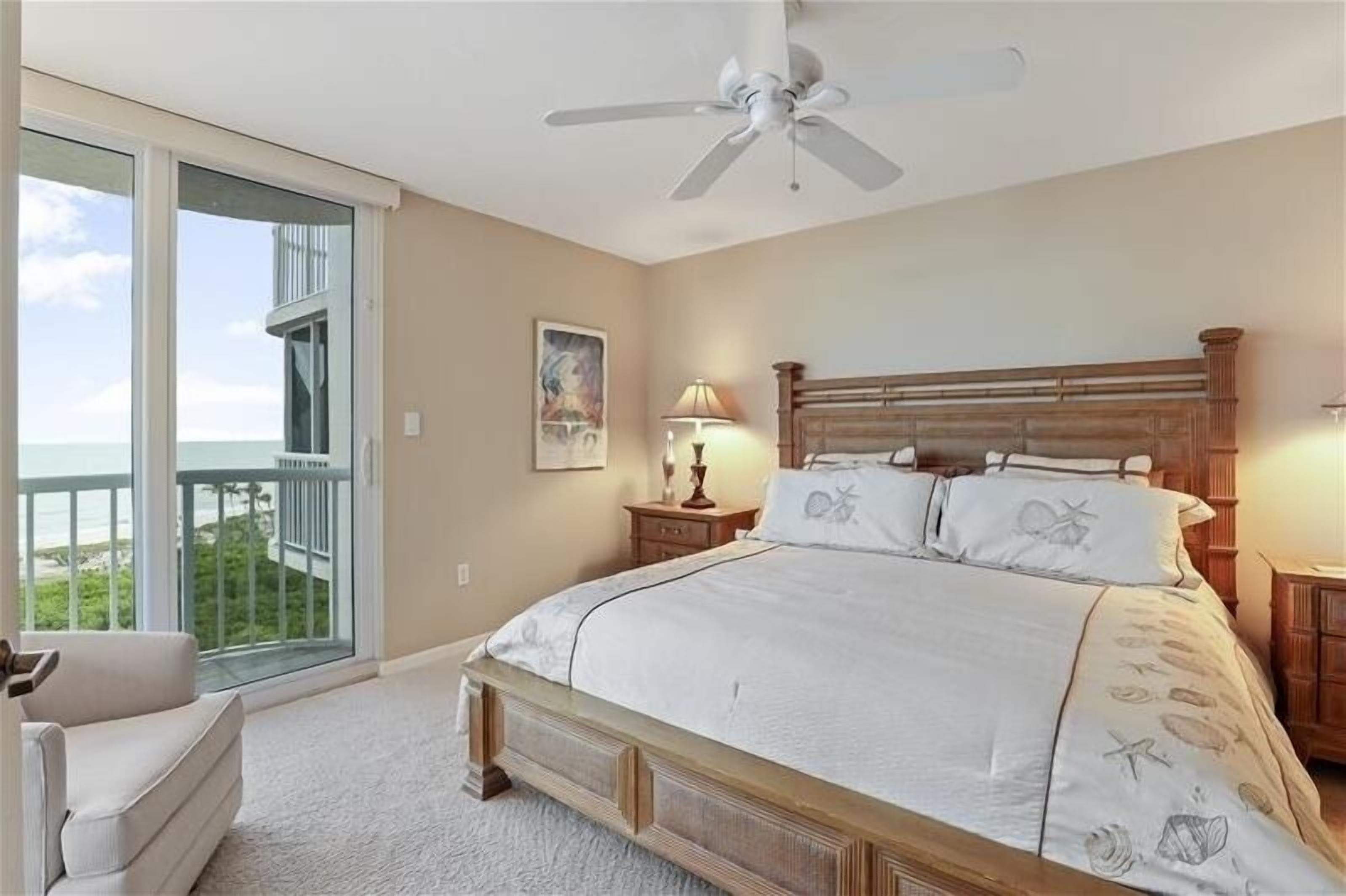 A bedroom with a large bed, a ceiling fan, a nightstand, a lamp, and a view of the ocean through a sliding glass door.