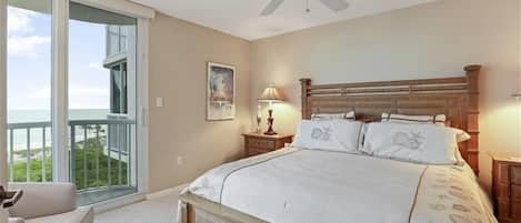 A bedroom with a large bed, a ceiling fan, a nightstand, a lamp, and a view of the ocean through a sliding glass door.