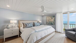 A bedroom with a large bed, a nightstand, a lamp, a painting, and a view of the ocean.