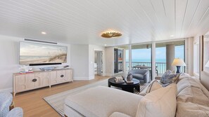 A modern living room with a large flat-screen TV, a sofa, a coffee table, and a view of the ocean through large windows.