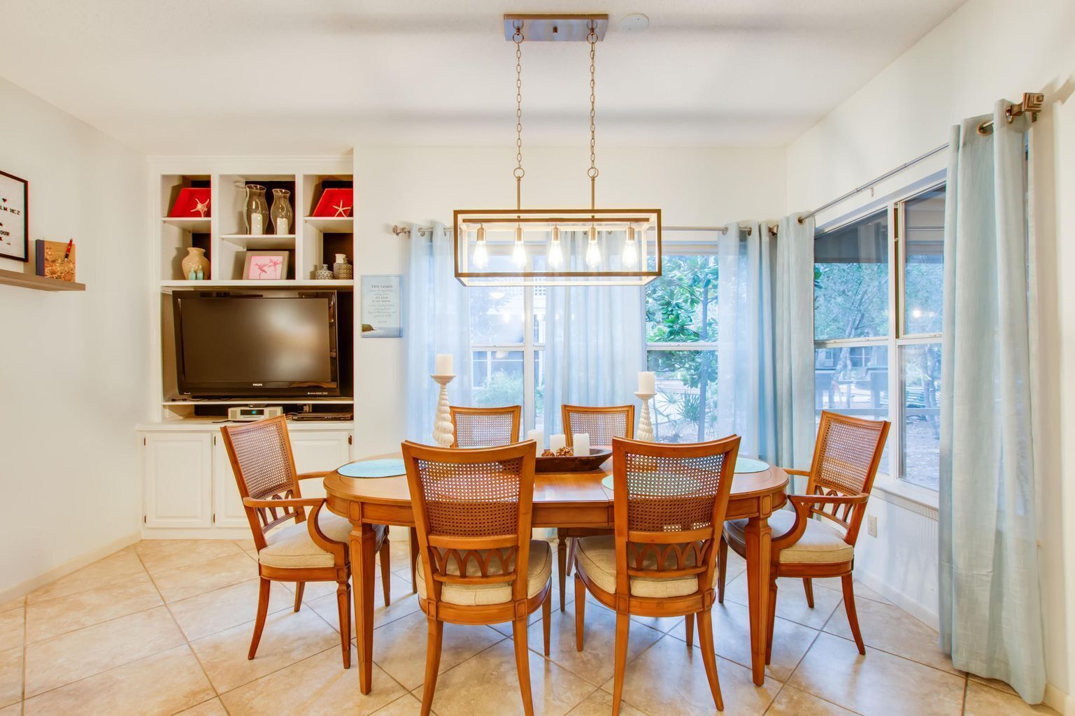 A dining area with a round table, six wooden chairs, a chandelier, a flat-screen TV on a white cabinet, and a window with curtains.