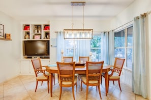 Uma sala de jantar com uma mesa redonda, seis cadeiras de madeira, um candelabro, uma televisão de tela plana sobre um armário branco e uma janela com cortinas.