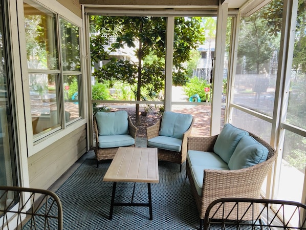 A covered patio with wicker furniture and a table, surrounded by large windows offering views of a garden and street.