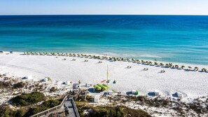A beach with white sand and clear blue water. There are rows of green umbrellas and lounge chairs. A wooden walkway leads to the beach.