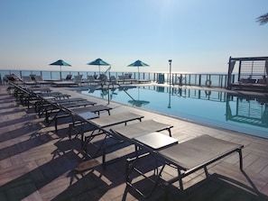 Área da piscina com espreguiçadeiras e guarda-sóis, céu limpo e vista para o mar.