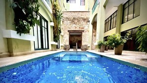 A swimming pool with a stone wall in the background, surrounded by potted plants and a building with large windows. - Iconic double apartment with billar in front of the Arco Chato ruins  and pool (Ciudad de Panamá)