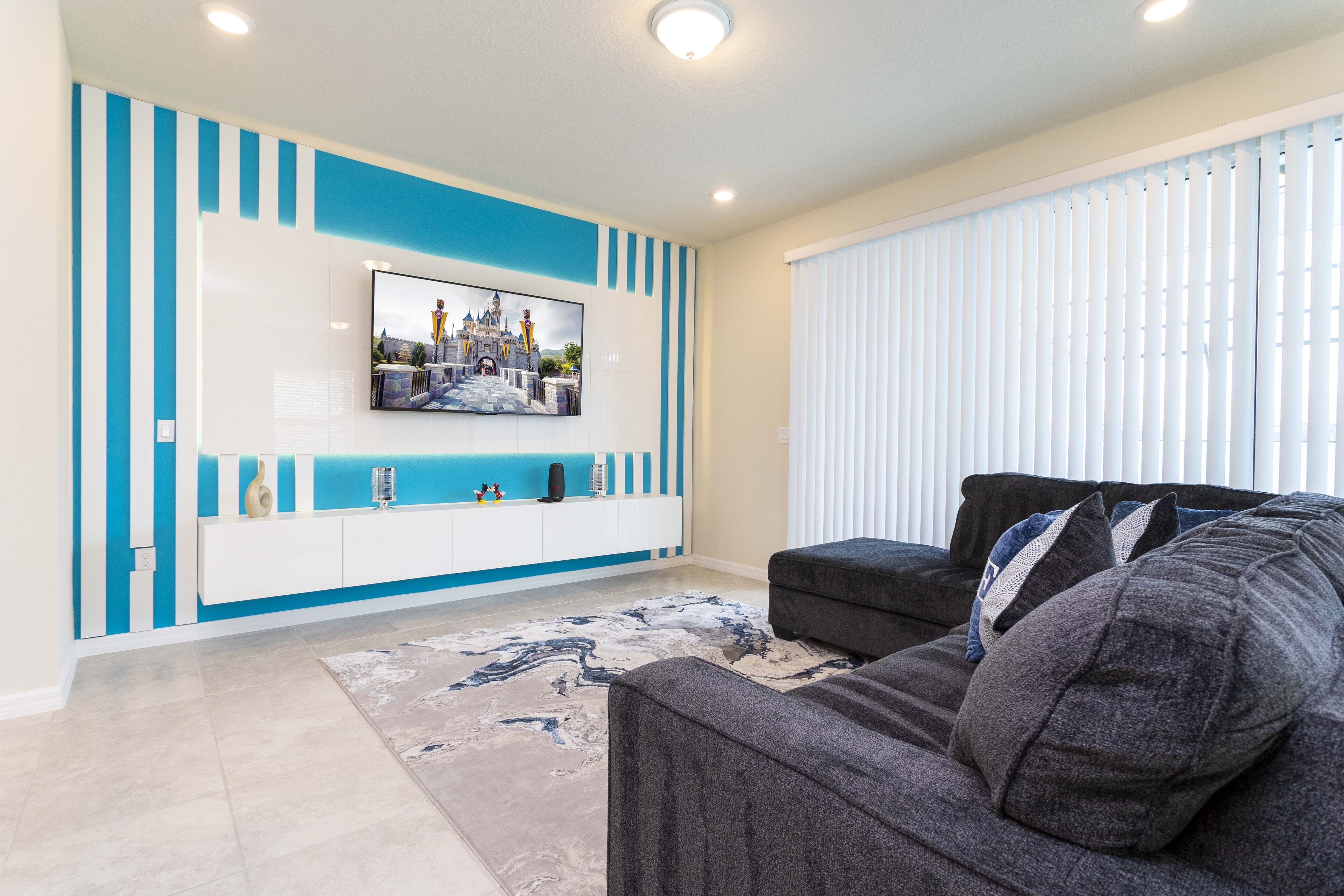 A modern living room with a flat-screen TV mounted on a white and blue wall unit, a dark grey sofa, and a patterned rug.