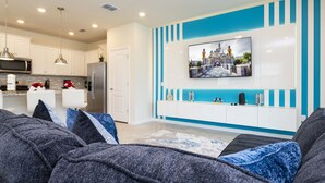 Un salon moderne doté d’un grand téléviseur à écran plat fixé au mur, d’un coin cuisine avec des meubles blancs et des appareils en acier inoxydable, et d’un espace salon confortable agrémenté de coussins gris et bleus.