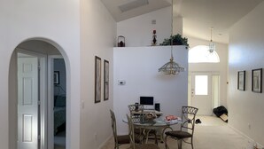 A dining area with a round table, chairs, and a chandelier. There is a door leading to another room and a plant on the wall.