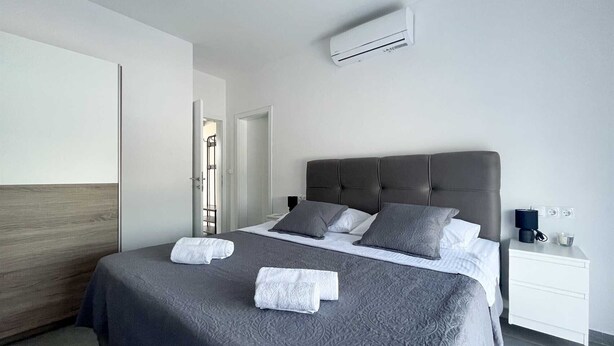 A modern bedroom with a grey upholstered bed, a white nightstand with a lamp, and an air conditioning unit above the bed.